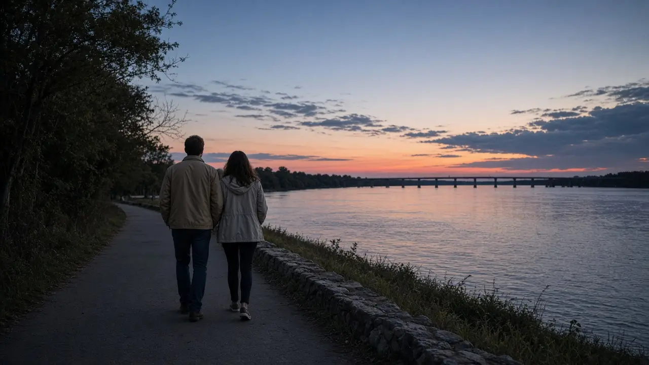 A dressed couple in light jackets on a riverside walk at dusk, a low bridge far away.