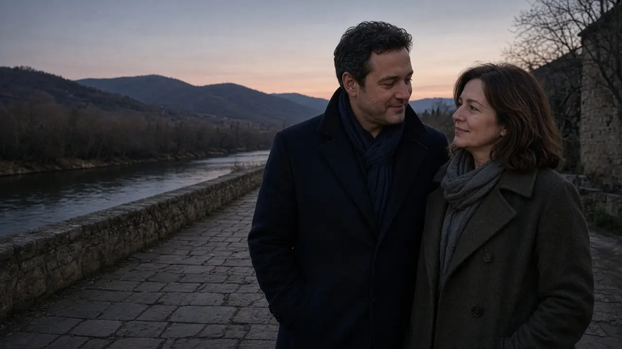 A dressed couple in coats on a stone riverside path at dusk, soft hills.