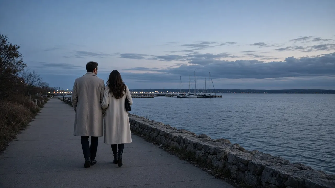 A dressed couple in light coats on a bayfront walk at dusk, sailboat masts far away, no signs.